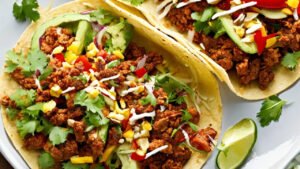 A white plate holds two tacos stuffed with ground beef and sautéed vegetables, garnished with lemon and cilantro.